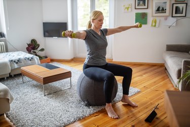 woman sitting down on a gray stability ball doing a dumbbell workout in living room
